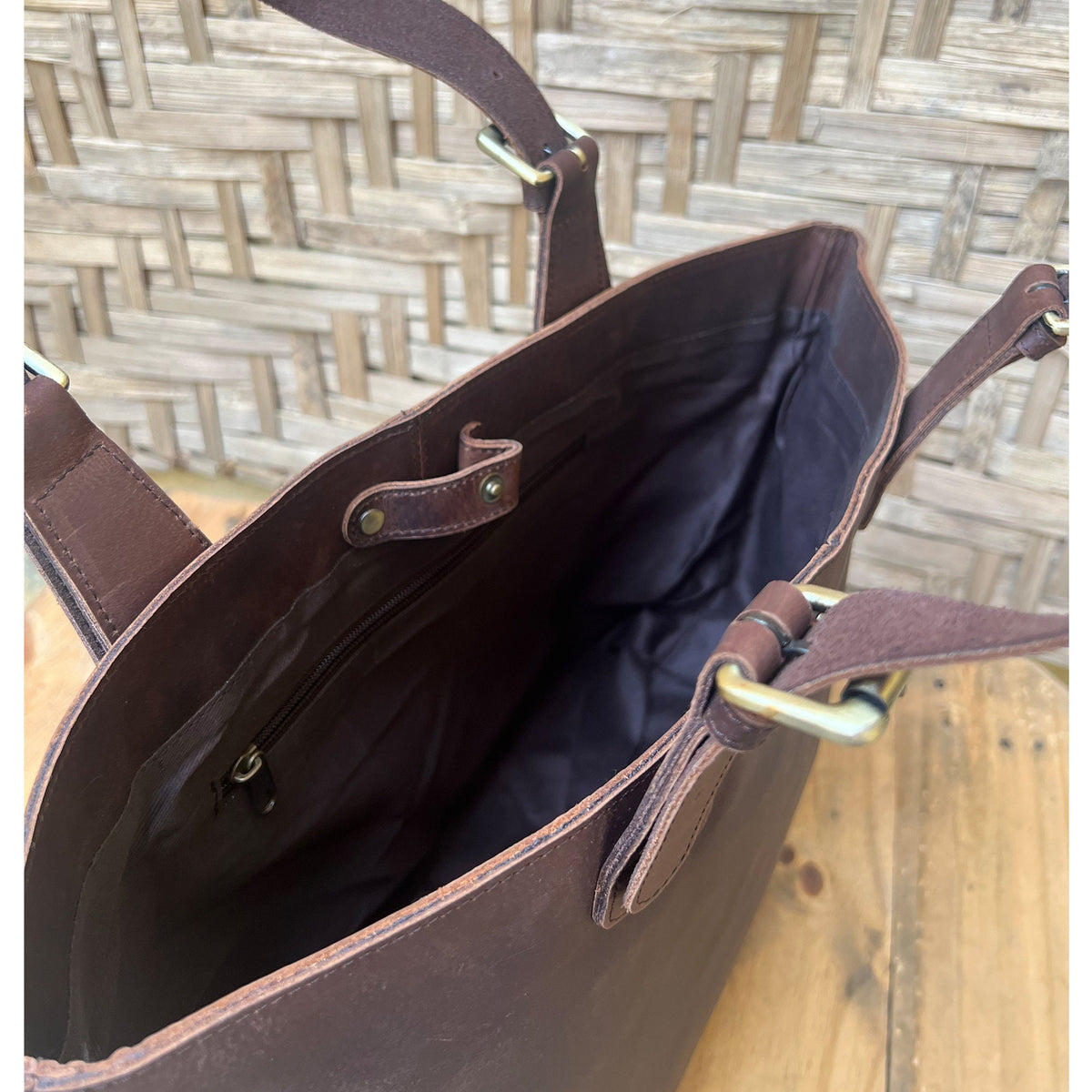 May include: A brown leather tote bag with a zippered pocket and two adjustable straps with gold buckles. The bag is open and the inside is lined with a dark fabric.