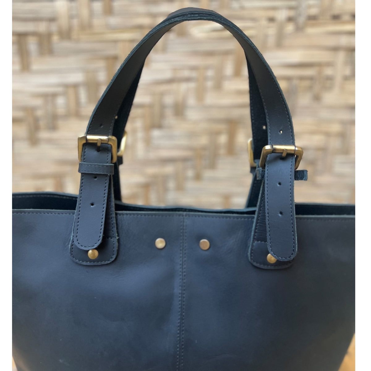May include: A dark blue leather tote bag with two adjustable straps and brass buckles. The bag has a simple design with a smooth leather finish.