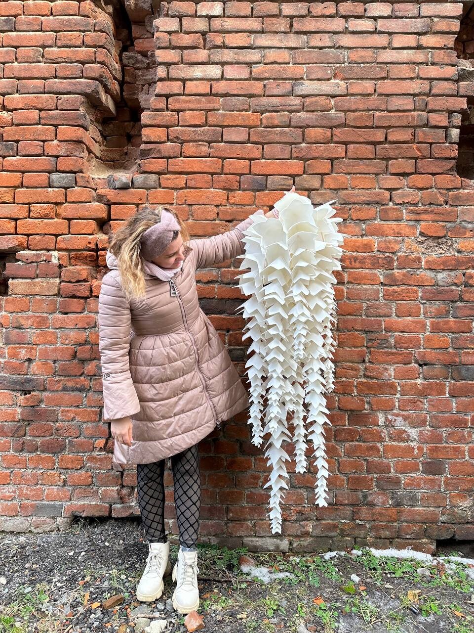 May include: A large, white, cascading paper sculpture with a layered, geometric design. The sculpture is held against a weathered brick wall. The piece has a unique, artistic aesthetic.