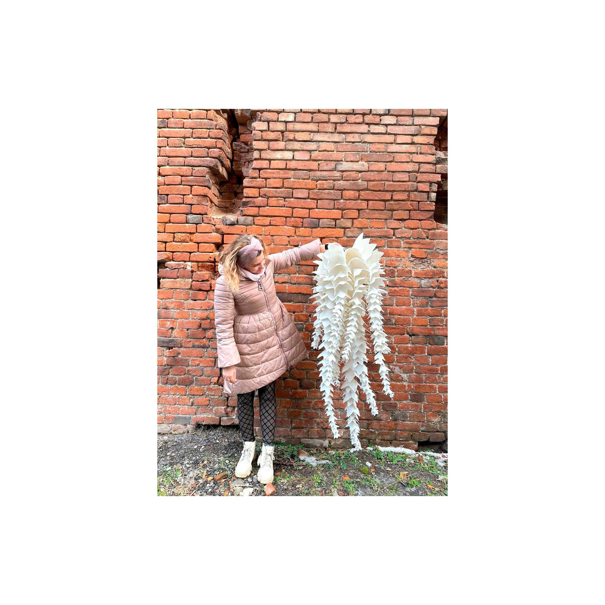 May include: A large, white, paper-like sculpture with cascading, fringed elements is held by a person wearing a pink coat. The sculpture is displayed against a weathered brick wall, creating a contrast of textures and colors. The piece appears to be an artistic, decorative item.
