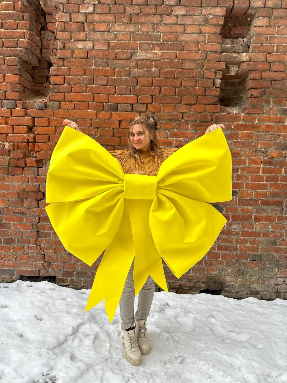May include: A woman is standing in front of a brick wall wearing a yellow bow. The bow is large and covers most of her body. She is smiling and looking at the camera.