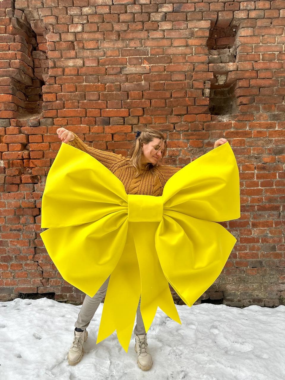 May include: A person wearing a brown sweater holds a large yellow bow in front of a brick wall. The bow is tied in a large knot and has long tails.