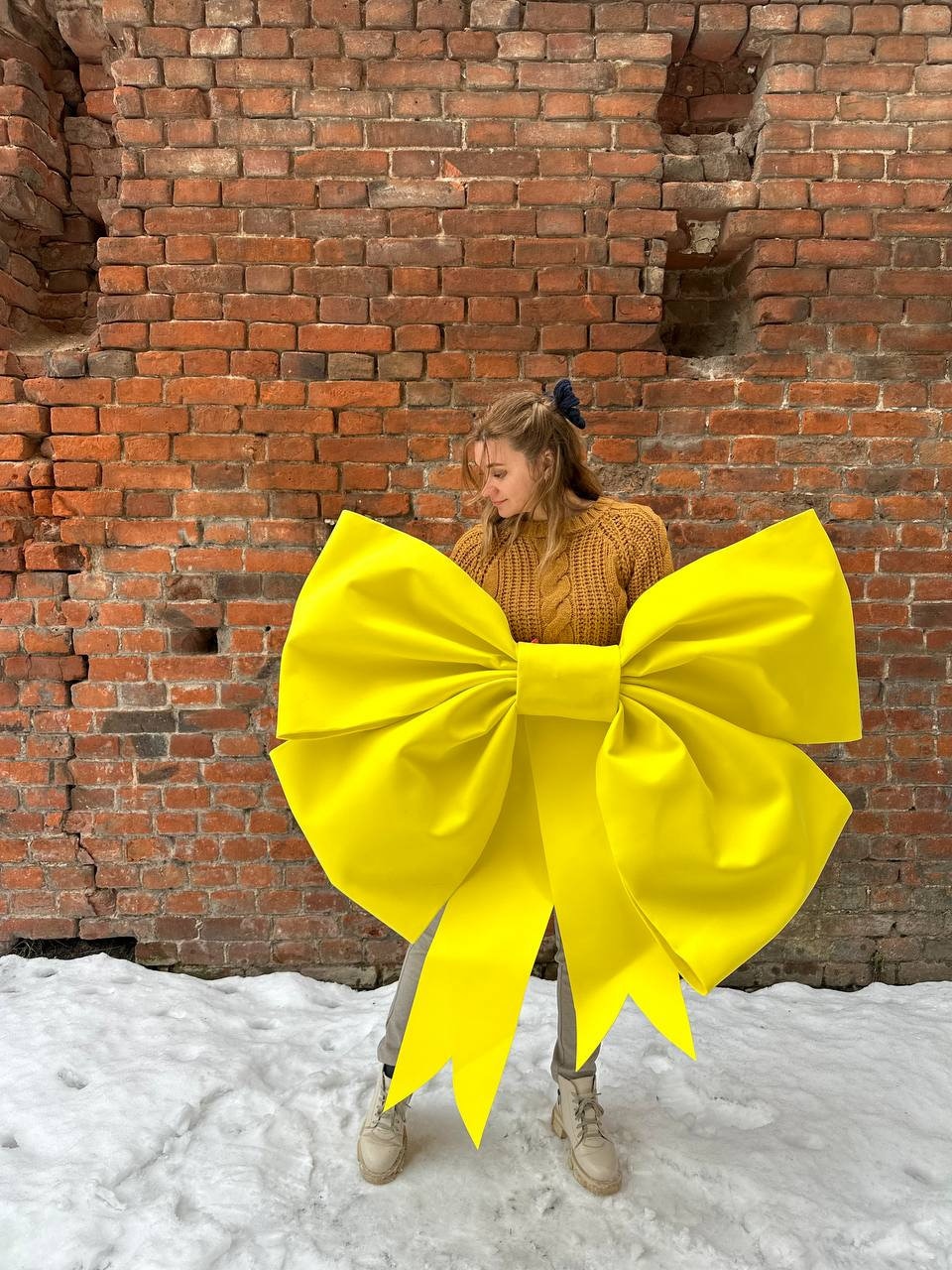 May include: A person wearing a brown sweater stands in front of a brick wall holding a large yellow bow. The bow is tied around the person's waist.