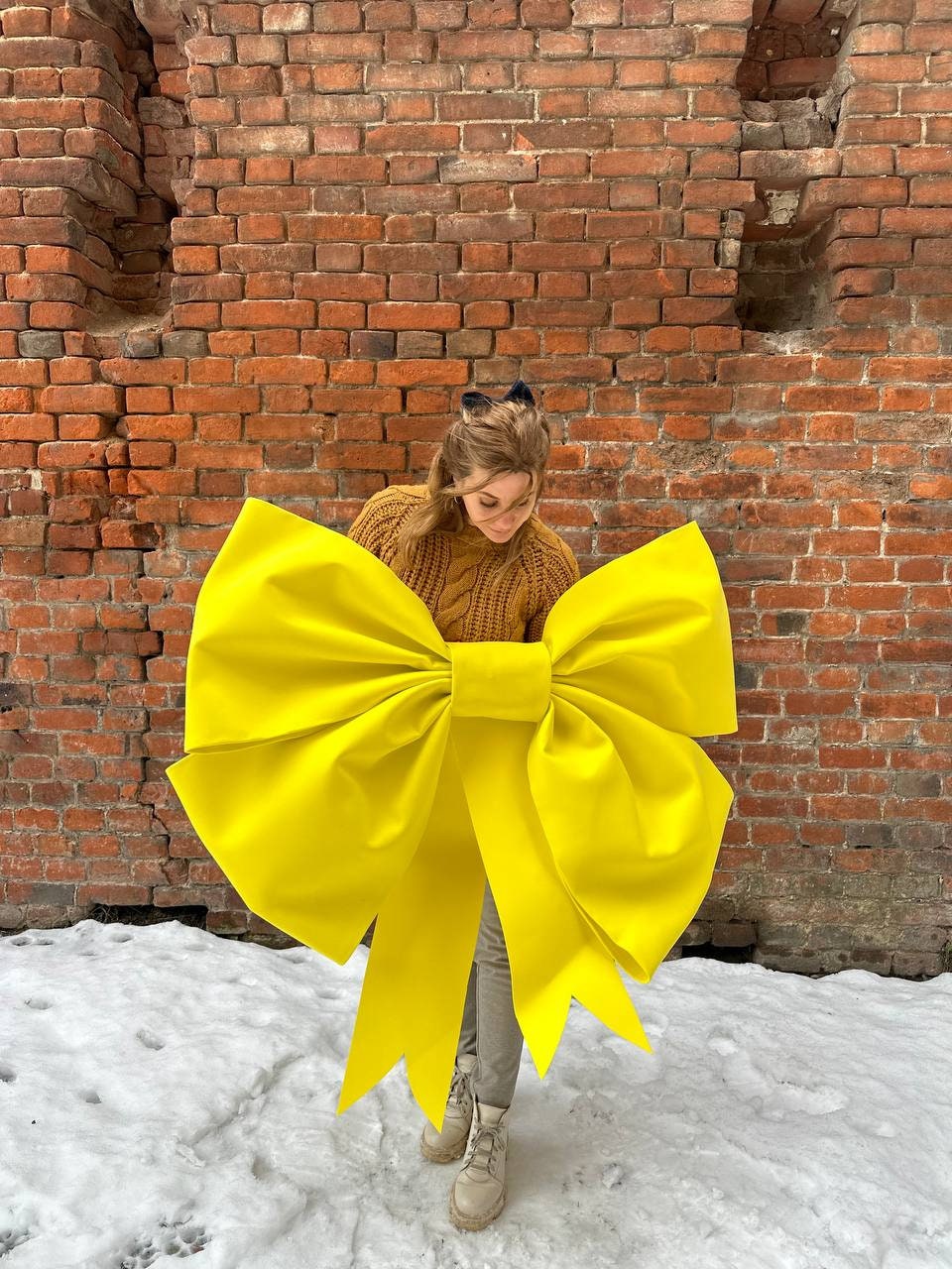 May include: A large, bright yellow bow is held in front of a person wearing a brown sweater. The bow is made of fabric and has a large, rounded center. The person is standing in front of a brick wall with snow on the ground.