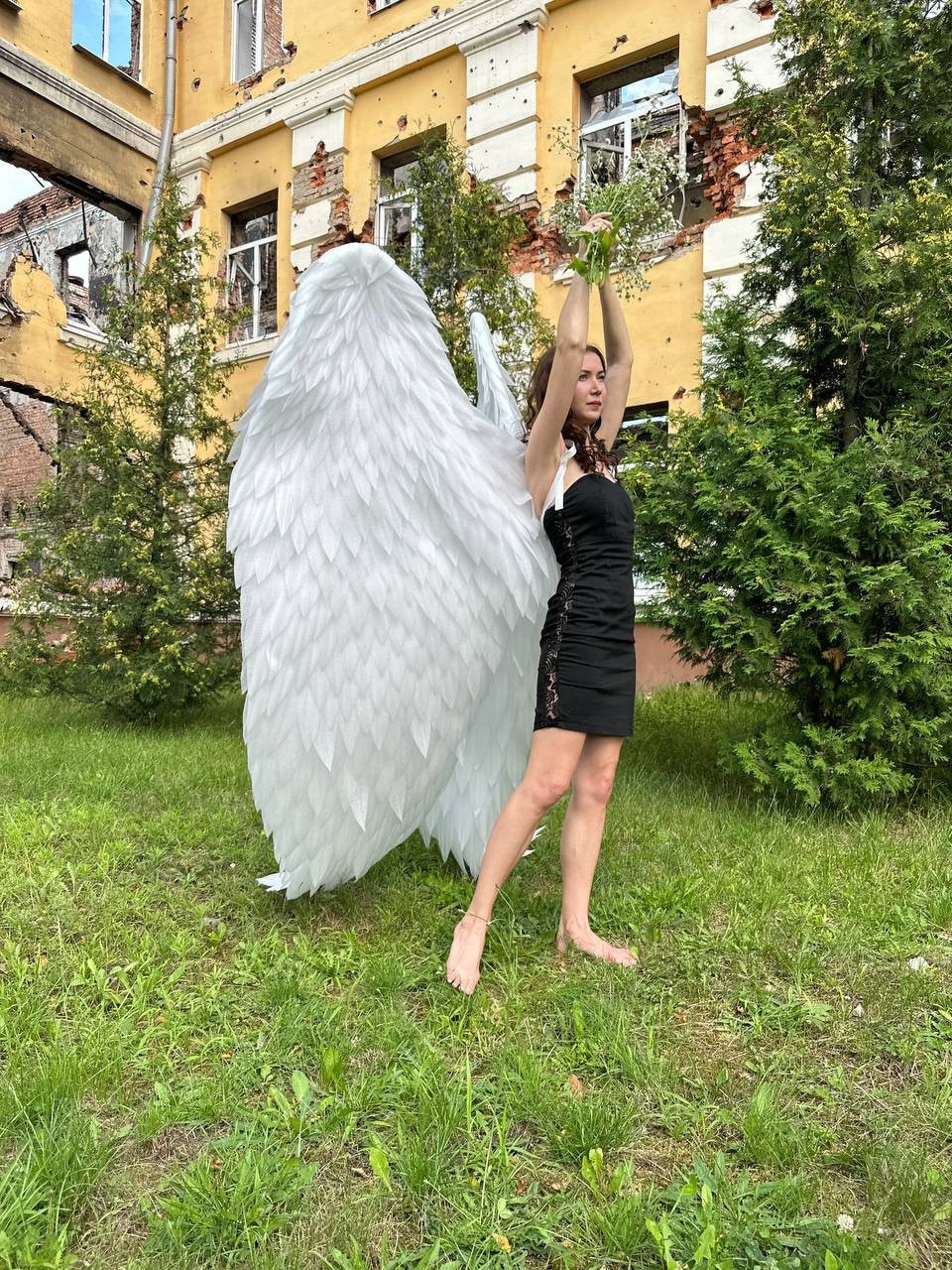 May include: A person wearing a black dress and large white angel wings stands in a grassy area in front of a building with bullet holes.