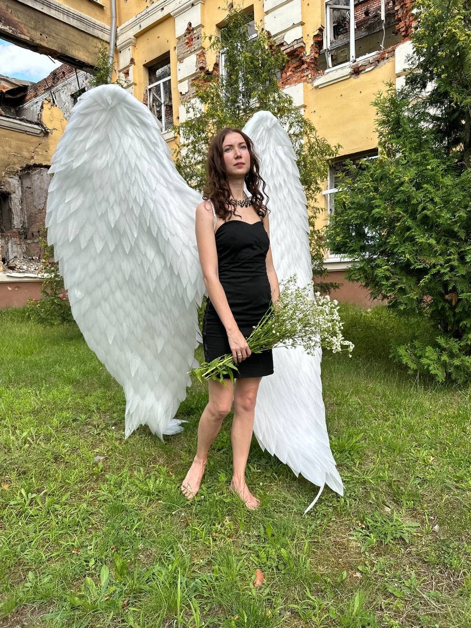 May include: A woman wearing a black strapless dress and white angel wings stands in a grassy area. She is holding a bouquet of white flowers and looking up.