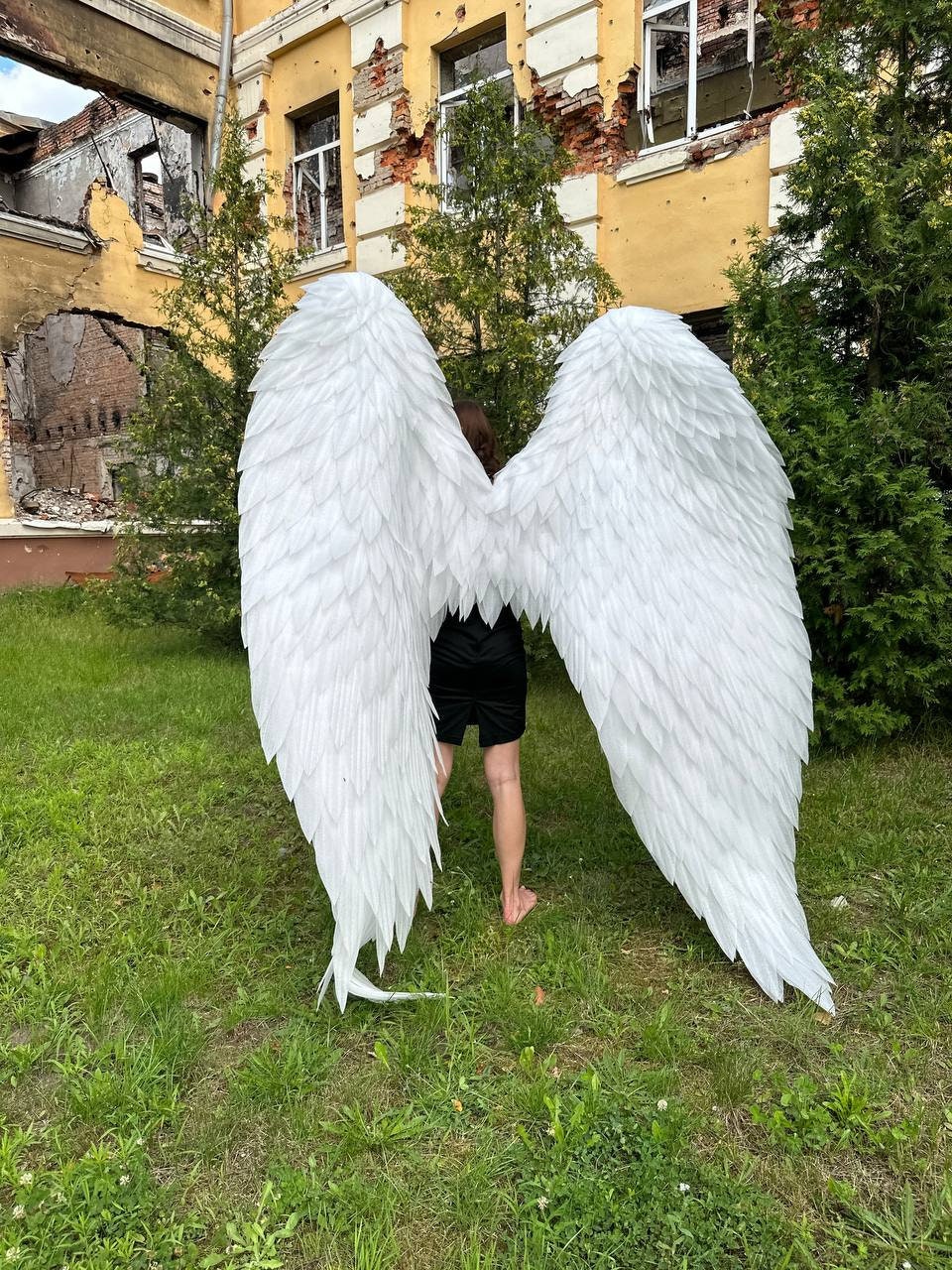 May include: A pair of large, white angel wings made of feathers. The wings are spread wide and are in front of a building with a damaged facade.