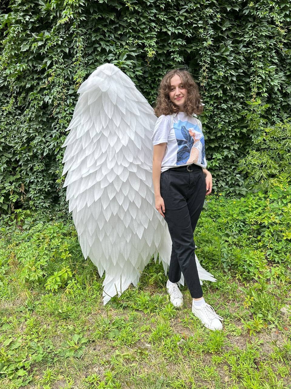 May include: A person standing in front of a large, white, paper angel wing sculpture. The wing is made of many small, overlapping paper feathers. The person is wearing a white t-shirt with a floral design and black pants.