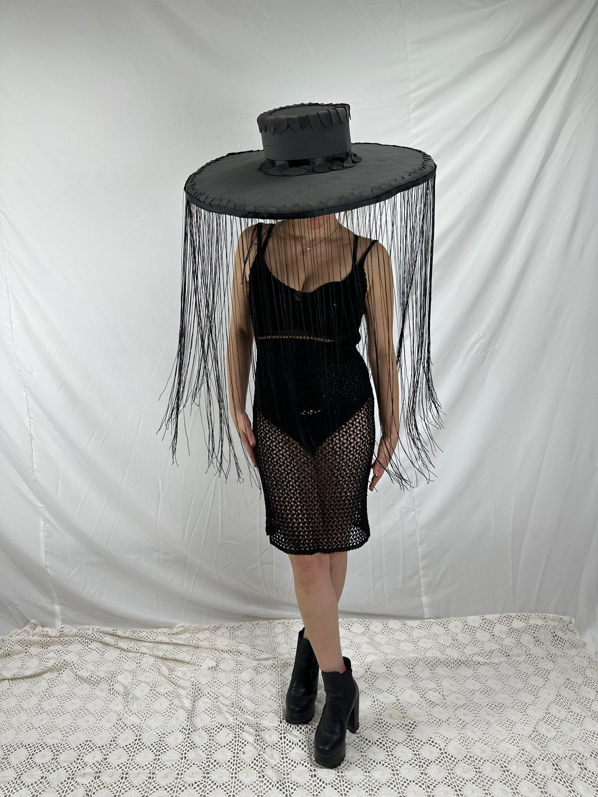 May include: A black wide-brimmed hat with a fringe trim. The hat is worn by a person wearing a black mesh dress and black boots.