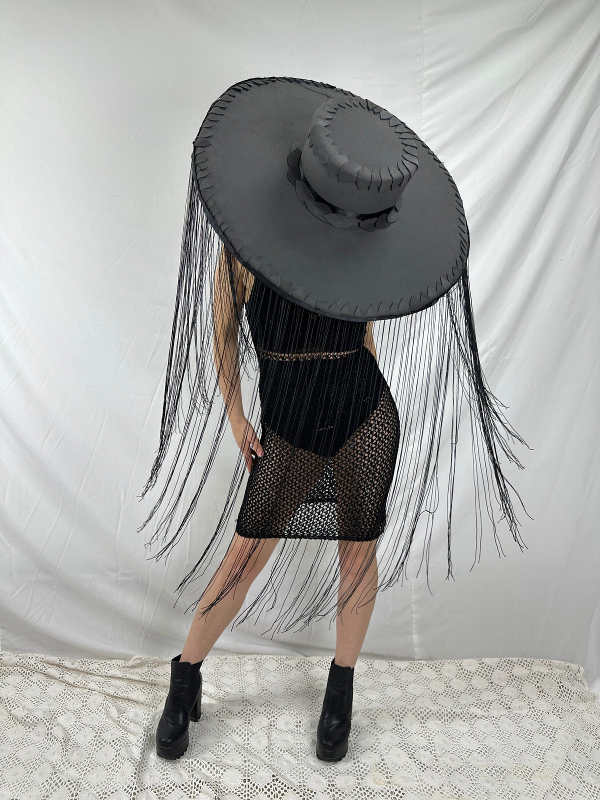 May include: A black wide-brimmed hat with fringe hanging from the brim. The hat is worn by a person wearing a black mesh dress and black boots.