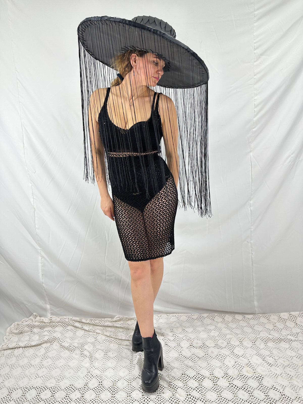 May include: A black wide-brimmed hat with long fringe hangs over a person wearing a black mesh dress and black heeled boots.