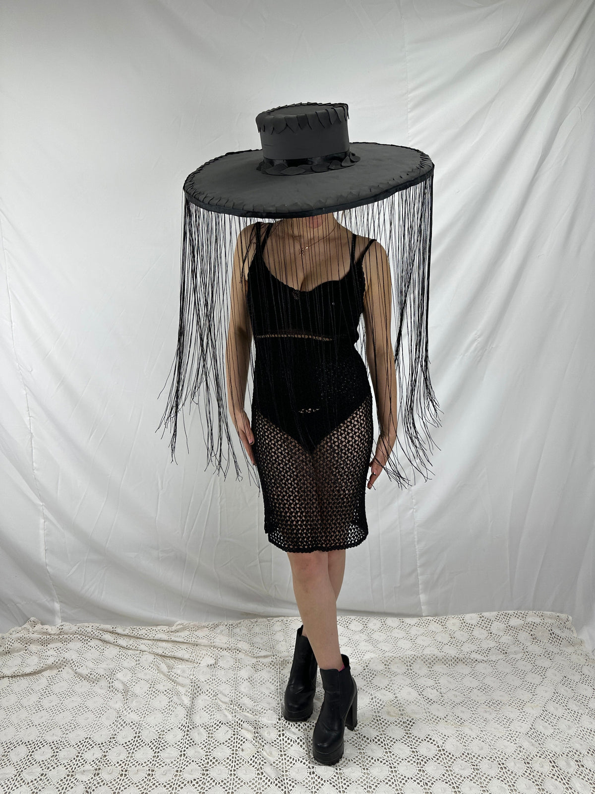 May include: A black hat with a wide brim and long fringe, paired with a black dress and platform boots. The hat's brim is approximately 24 inches in diameter. The dress is a crochet style, and the boots have a chunky heel.