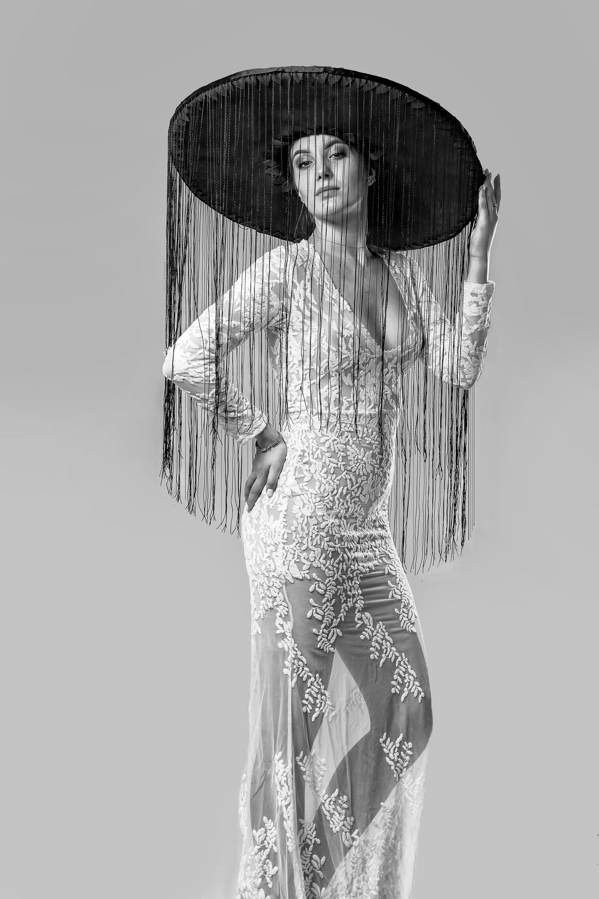 May include: A black and white image of a woman wearing a large, wide-brimmed hat with long fringe. She is also wearing a long, white, lacy dress with a plunging neckline. The dress has a floral pattern. The overall aesthetic is elegant and dramatic.