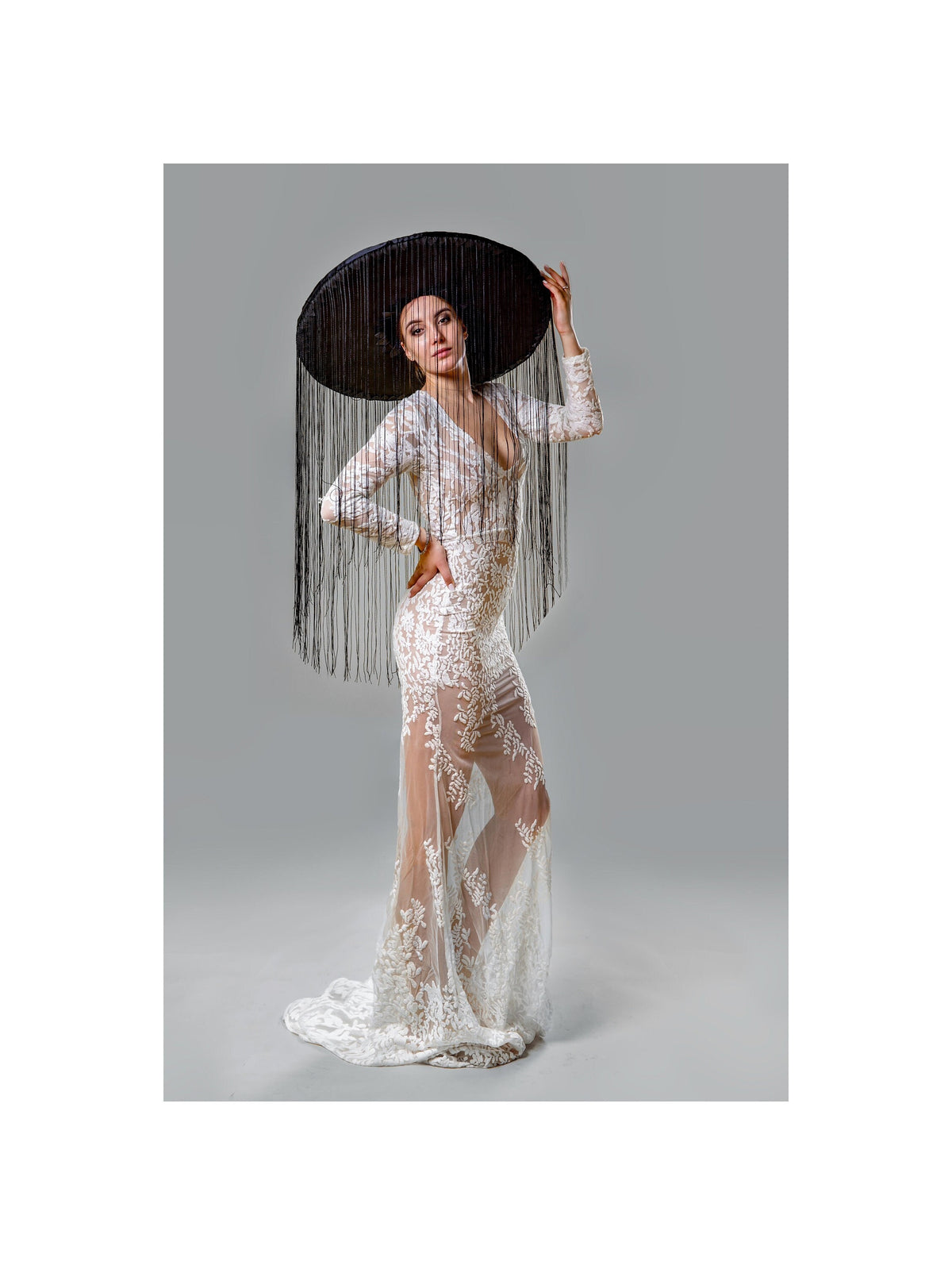 May include: A woman wearing a white lace gown with long sleeves and a plunging neckline. She is wearing a large black hat with long fringe. The dress has a sheer skirt and a train.