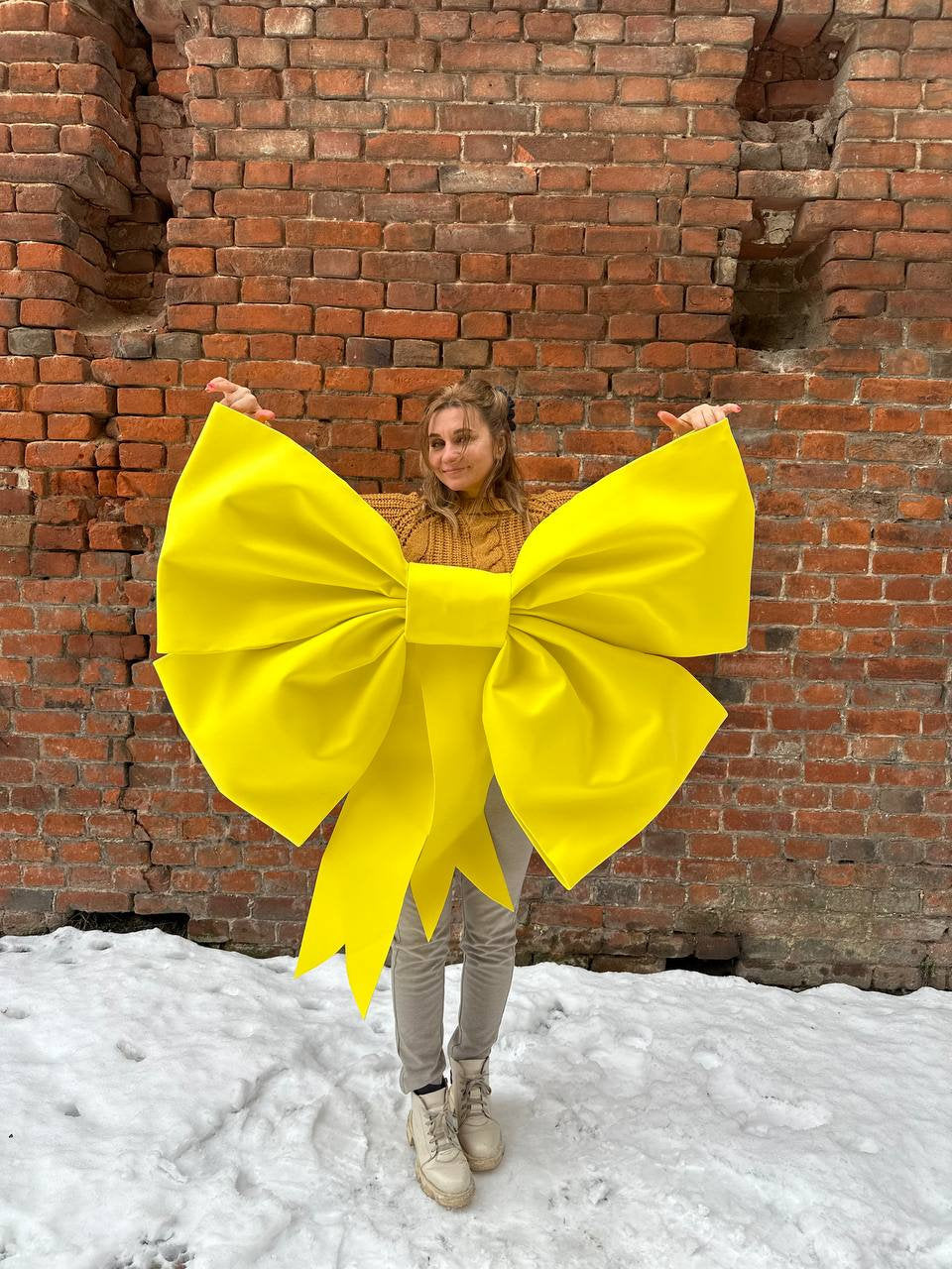May include: A person wearing a large, bright yellow bow made of fabric. The bow is tied around the person's waist and the ends of the bow are hanging down. The person is standing in front of a brick wall with snow on the ground.