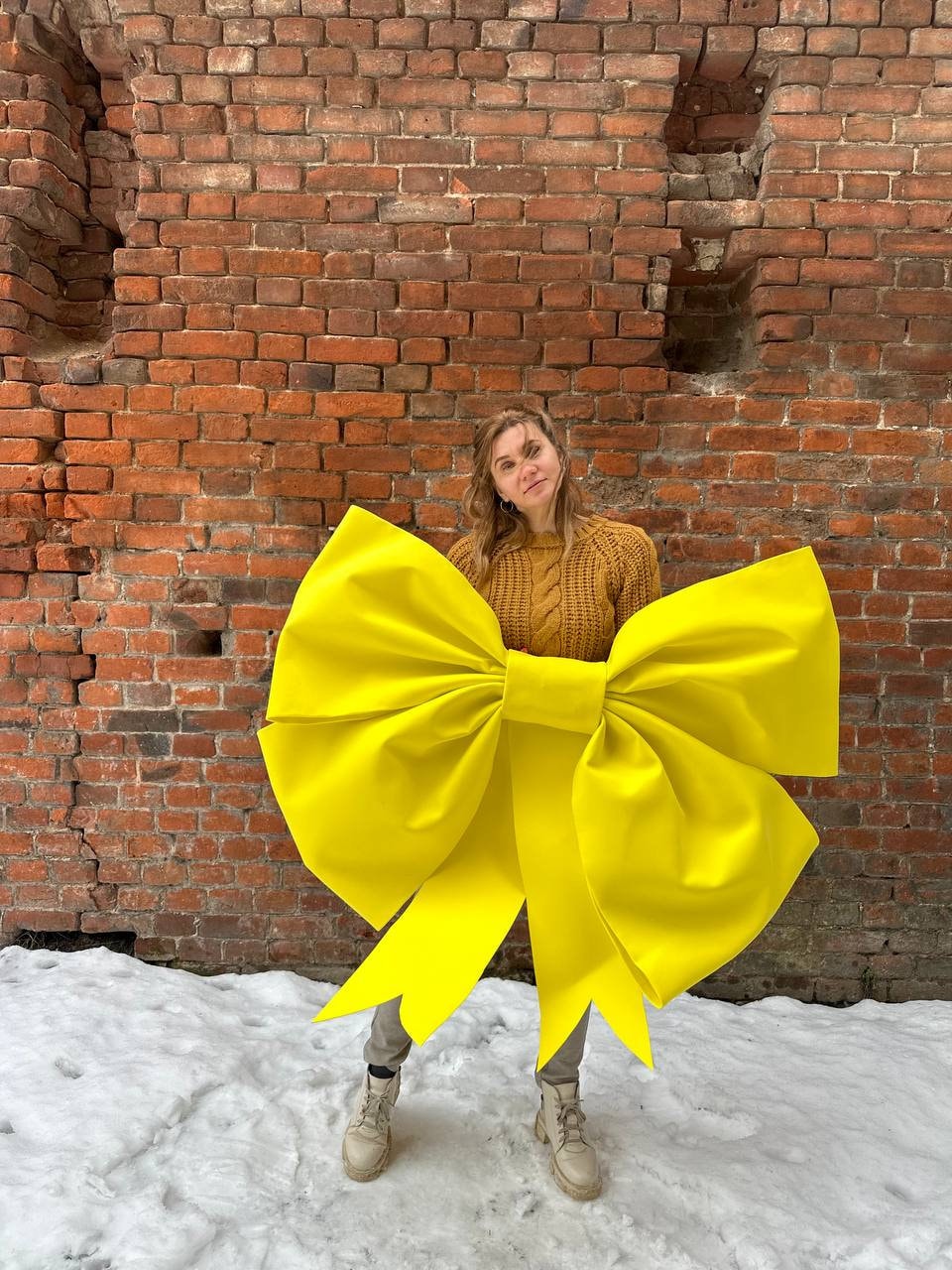 May include: A person is holding a large, bright yellow bow in front of a brick wall. The bow is made of fabric and has a large, central knot. The person is wearing a brown sweater and jeans.