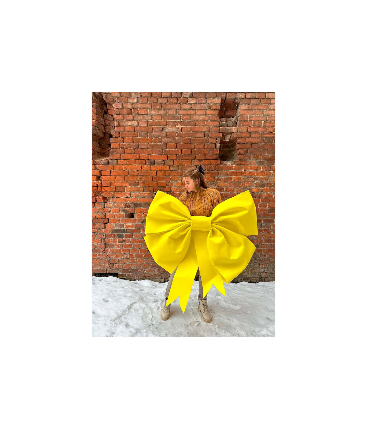 May include: A large yellow bow, tied in a classic shape, is held by a person in front of a brick wall. The bow is made of a shiny, smooth material and has a large, central loop.