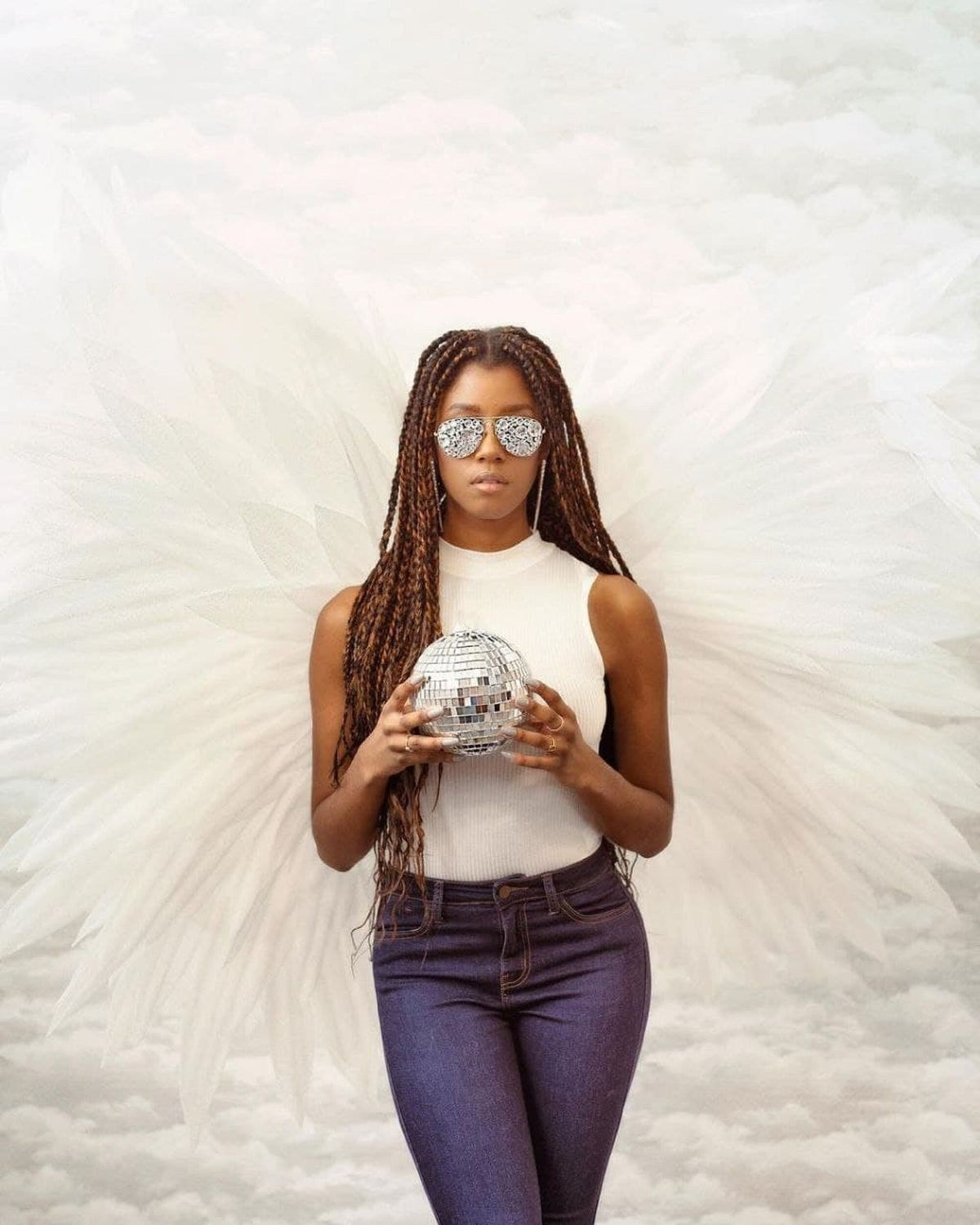 May include: A woman with long brown braids wears mirrored sunglasses and a white tank top. She holds a silver disco ball in front of her. She is standing in front of a white background with a white feathery angel wing on each side.