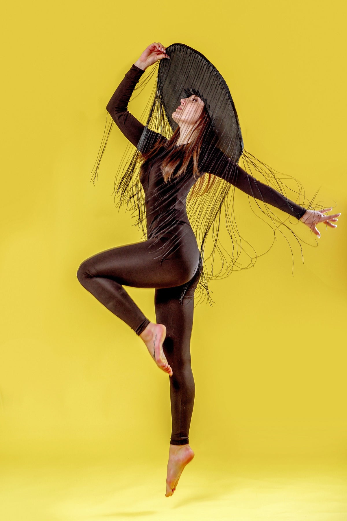 May include: A woman wearing a black jumpsuit and a large black hat with fringe, posing against a yellow background.