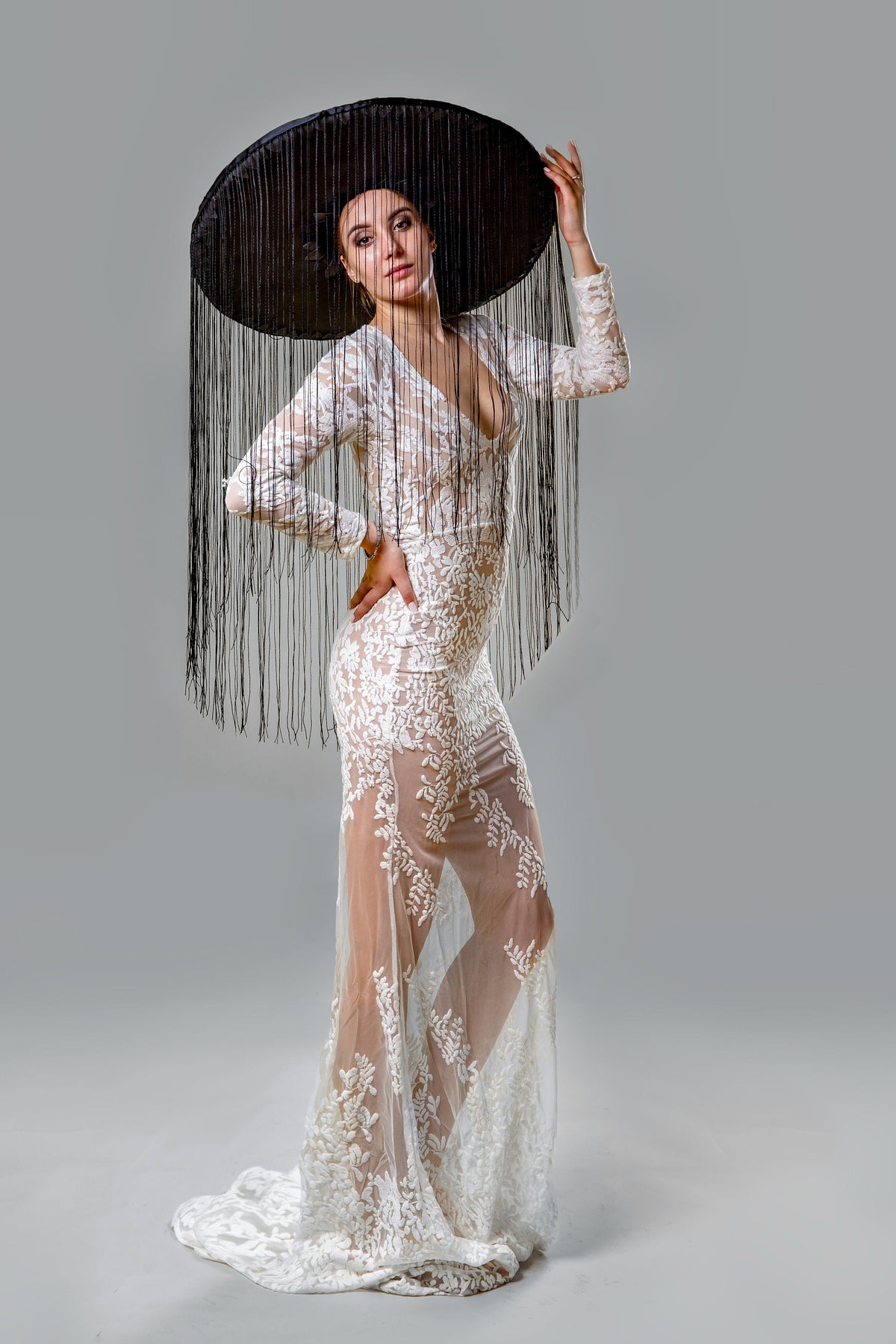 May include: A woman wearing a white lace gown with a plunging neckline and long sleeves. She is wearing a large black hat with long black fringe. The dress has a sheer skirt with floral embroidery.