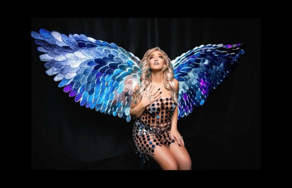 May include: A woman with long blonde hair and a sequined dress poses in front of large, blue and purple angel wings. The wings are detailed with layered, scale-like patterns, creating a dramatic effect against the black background.