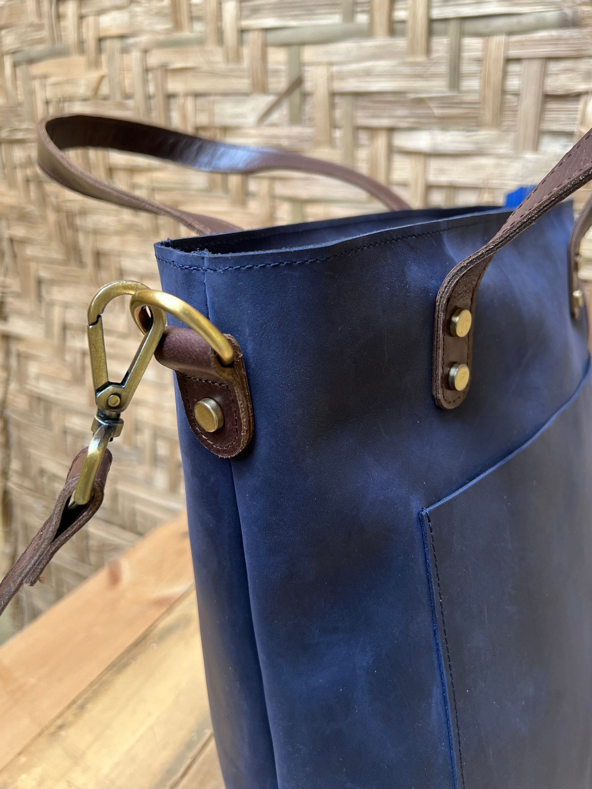 May include: A dark blue leather tote bag with brown leather straps and a brass hook. The bag has a front pocket and a top zipper closure.