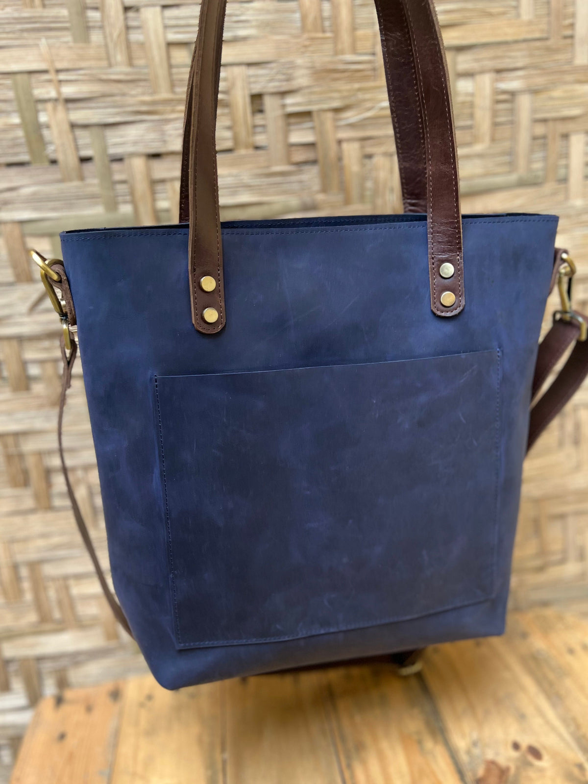 May include: A dark blue leather tote bag with brown leather straps and a front pocket. The bag has gold metal accents.