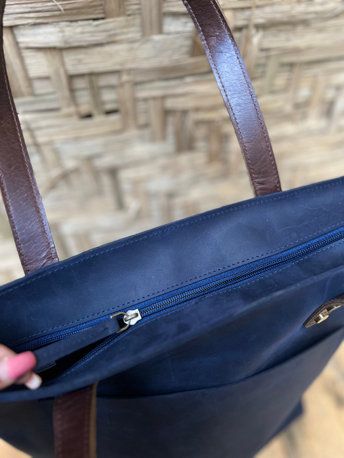 May include: A navy blue tote bag with brown leather straps and a zipper closure. The bag has a pocket on the front.
