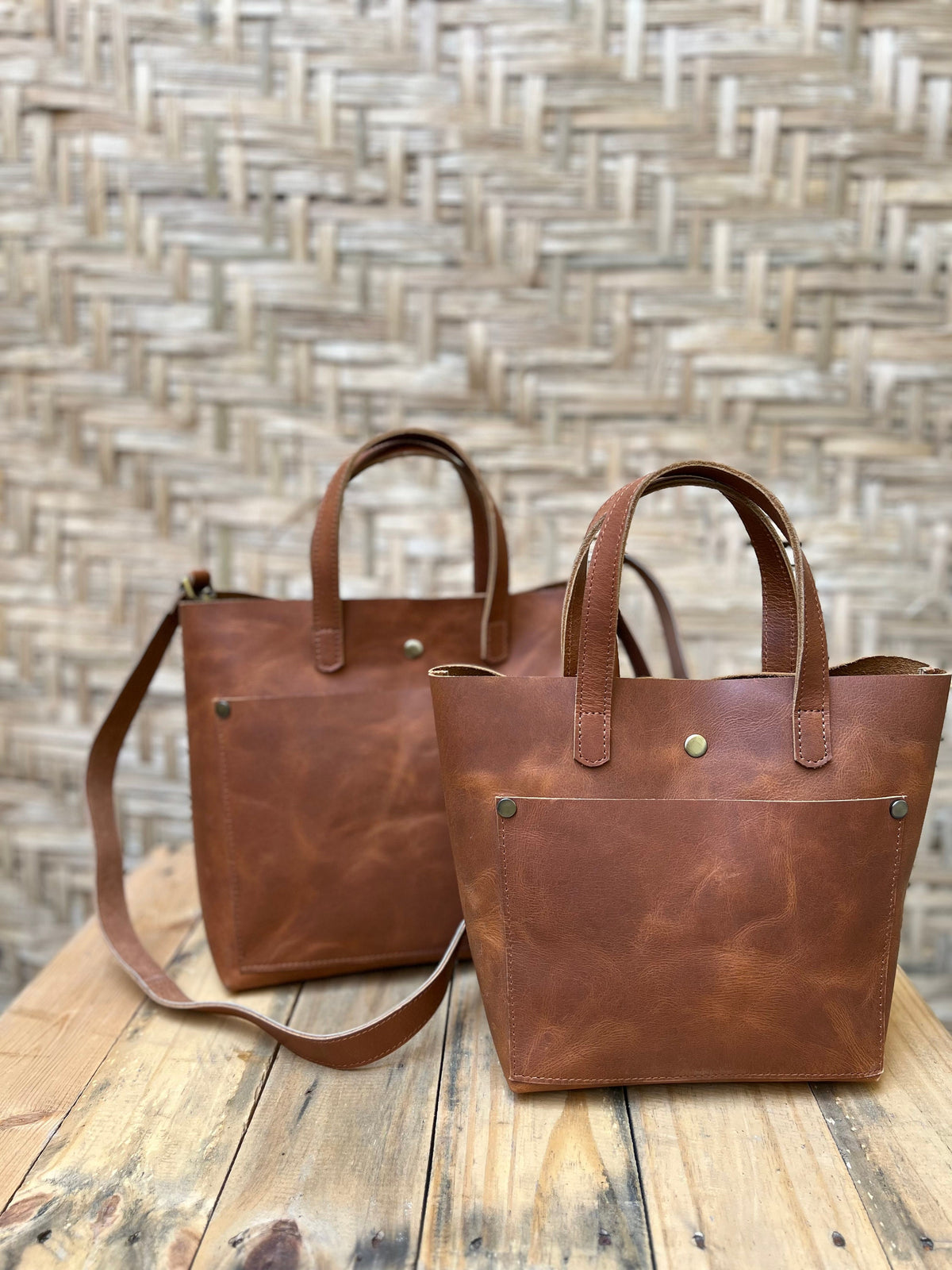 May include: Two brown leather tote bags with a single strap on the larger bag. The bags have a pocket on the front and a brass button closure.