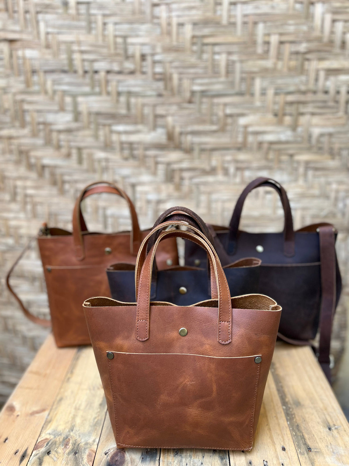 May include: Three brown leather tote bags with a front pocket. The bags have a top handle and a shoulder strap. The bags are arranged in a row on a wooden surface.