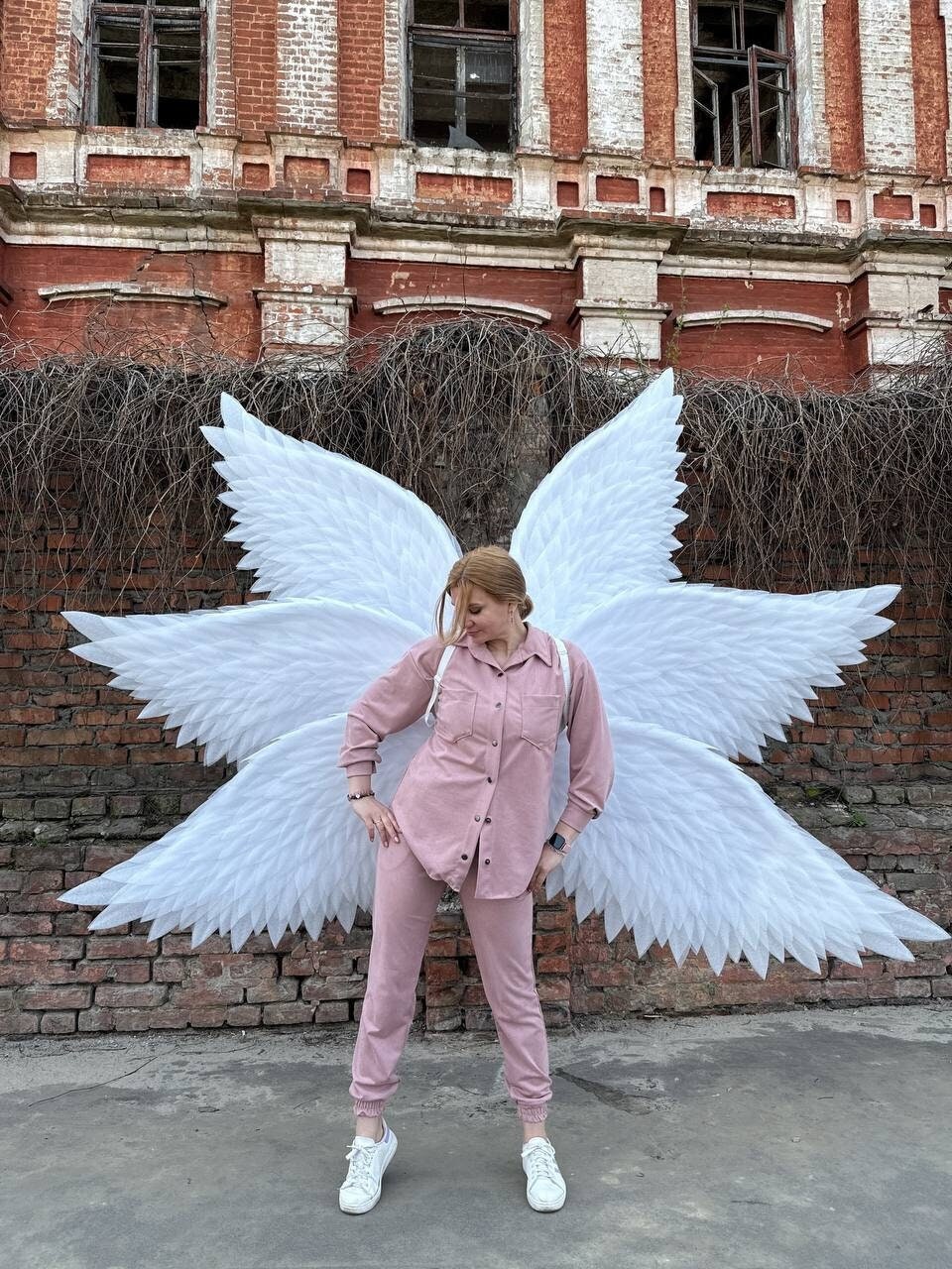May include: A person wearing a pink shirt and pants stands in front of a brick wall with large white angel wings behind them.