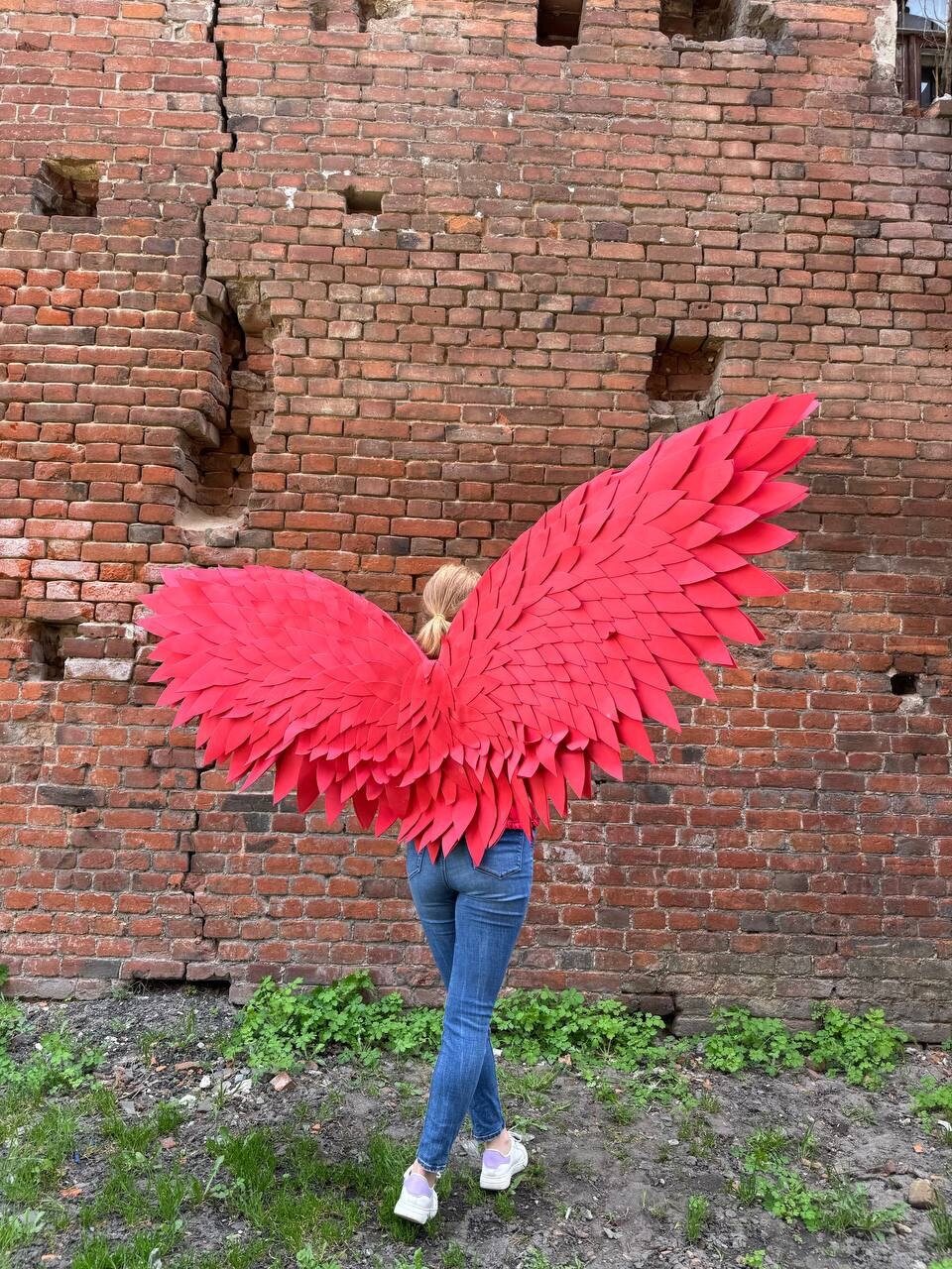 May include: A person wearing a pair of large, red, paper wings. The wings are made of many individual pieces of paper that are layered and overlapping. The person is standing in front of a brick wall.