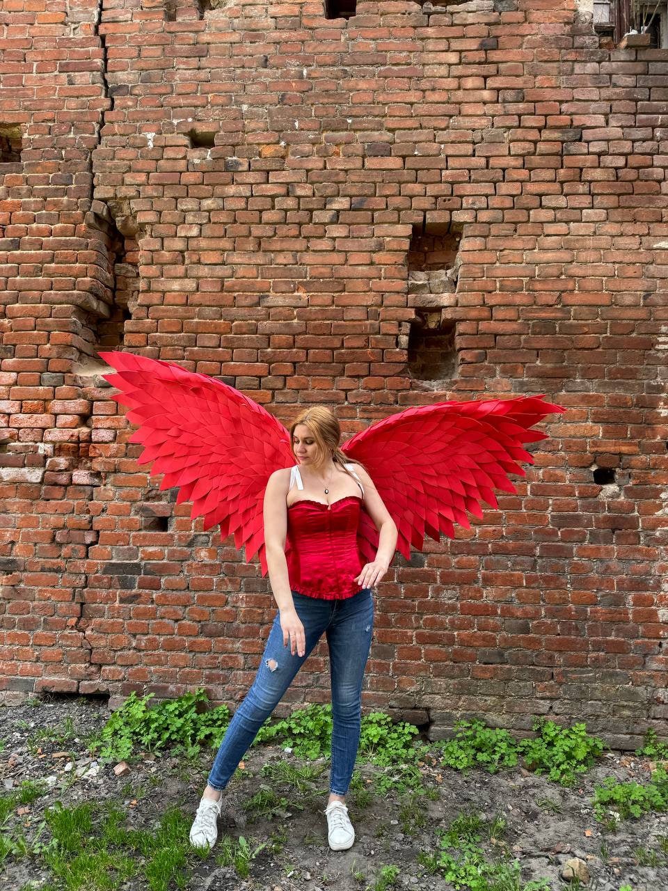 May include: A person wearing a red corset and blue jeans with large red feathered wings behind them. The person is standing in front of a brick wall.
