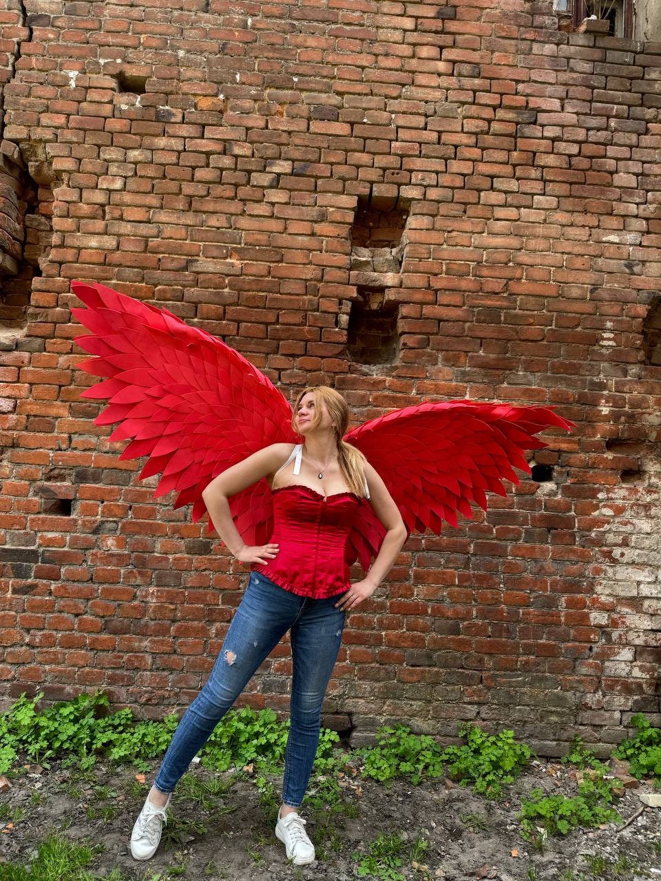 May include: A woman wearing a red corset and blue jeans with large red feathered wings behind her. She is standing in front of a brick wall.