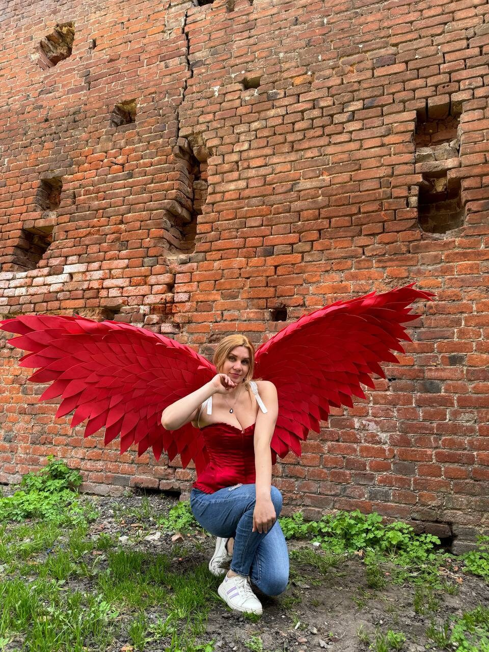 May include: A woman wearing a red corset and blue jeans poses with large red feathered wings in front of a brick wall.