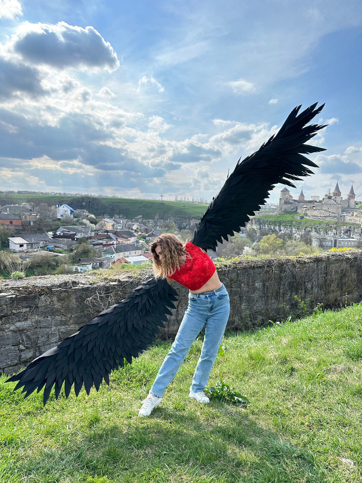 May include: A person wearing a red crop top and blue jeans is holding large black feathered wings. The wings are spread wide and the person is standing on a grassy hill with a stone wall behind them.
