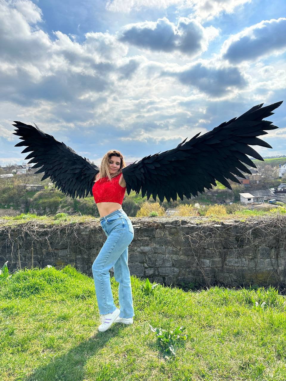 May include: A person wearing a red crop top and blue jeans is standing in a grassy field with a pair of large black wings behind them. The wings are made of feathers and are spread out wide. The sky is blue with white clouds.