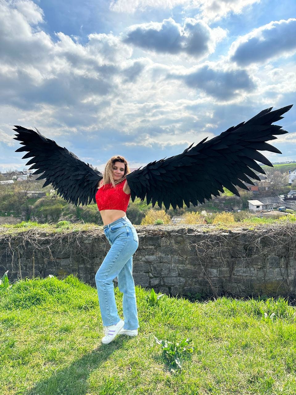 May include: A person wearing a red crop top and light blue jeans stands in a grassy area, with large black angel wings spread behind them. The wings are detailed and textured, set against a cloudy sky.