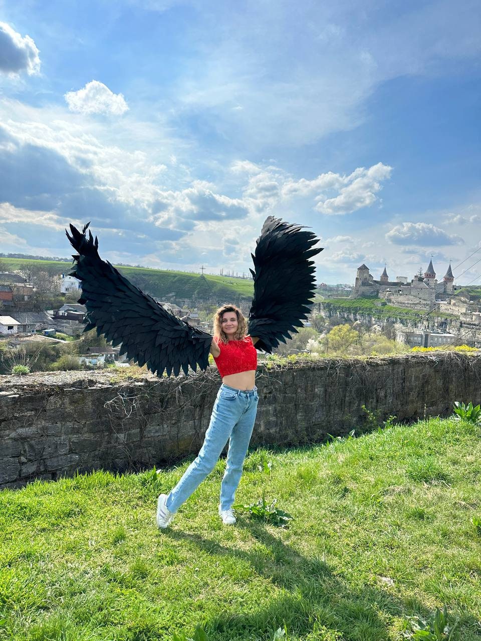 May include: A person wearing a red crop top and blue jeans is holding large black angel wings in front of a castle and a blue sky with white clouds.
