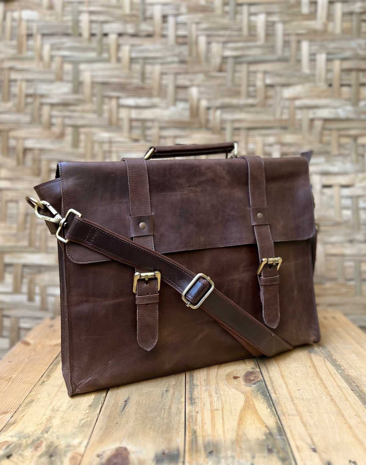 May include: A brown leather briefcase with a top handle and adjustable shoulder strap. The bag features two front straps with brass buckles and a flap closure. The briefcase is sitting on a wooden surface.