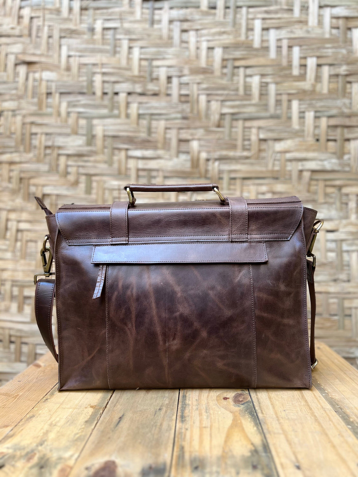May include: A brown leather briefcase with a top handle and a shoulder strap. The bag has a flap closure and brass-colored hardware. The leather has a distressed appearance, and the bag sits on a wooden surface.