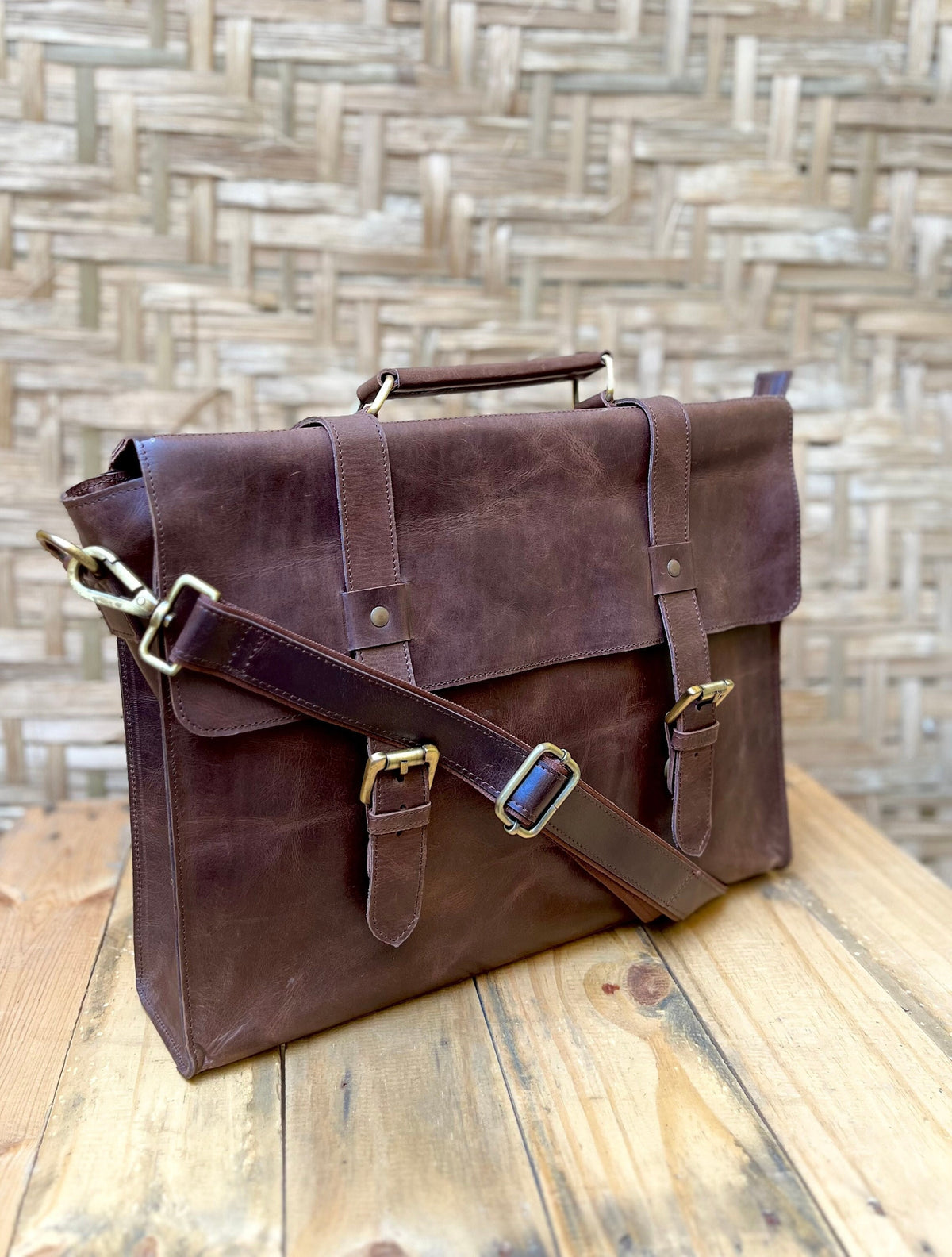 May include: A dark brown leather briefcase with a top handle and adjustable shoulder strap. The bag features brass-colored hardware, including buckles and a clasp. The briefcase is sitting on a wooden surface.