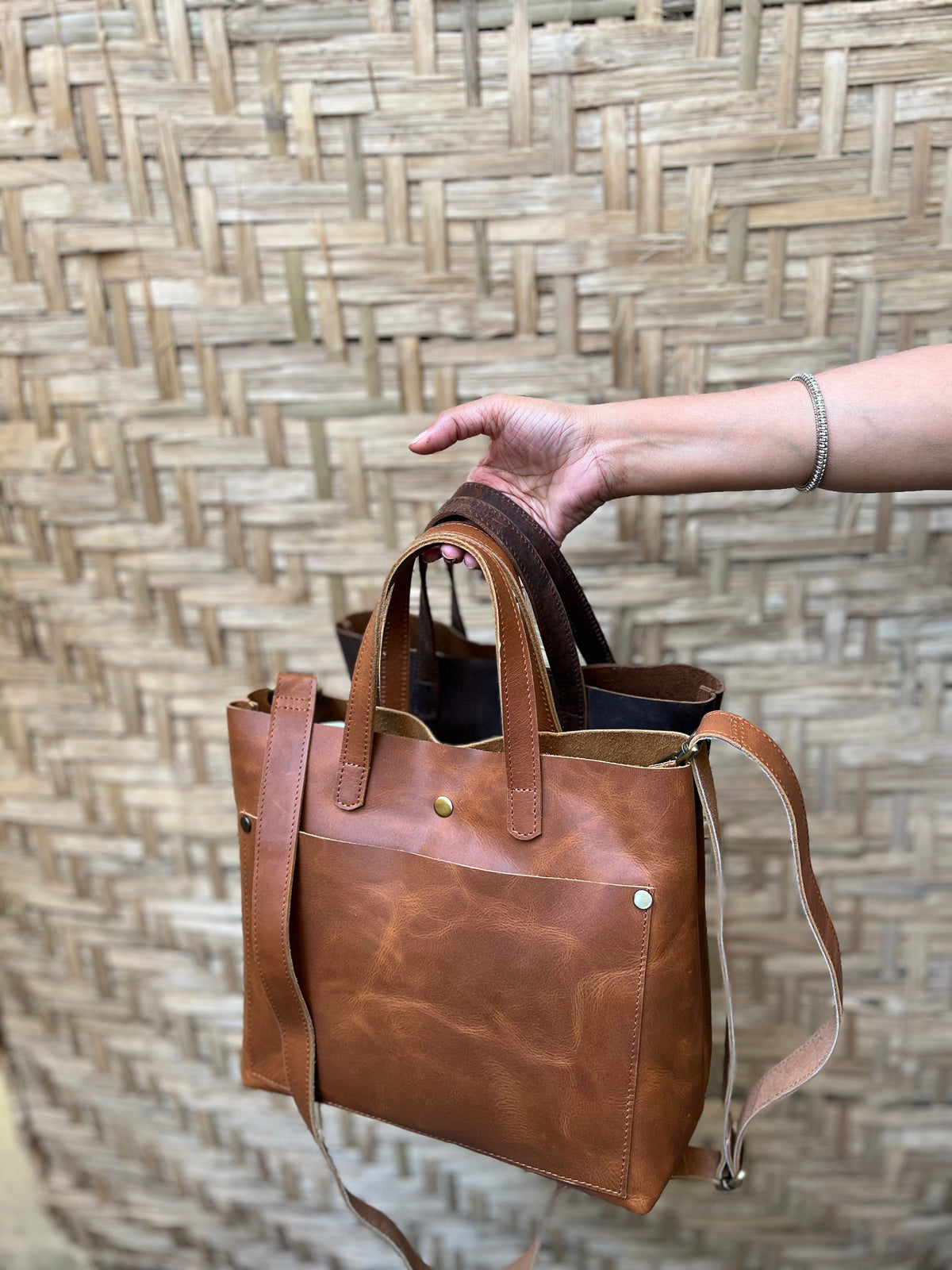 May include: A brown leather tote bag with a long strap and a front pocket. The bag has a distressed look and is made of high-quality leather.
