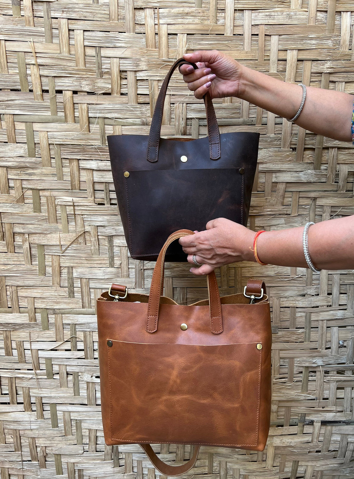 May include: Two leather tote bags, one brown and one tan, with a front pocket and a strap. The bags are displayed against a woven bamboo background.