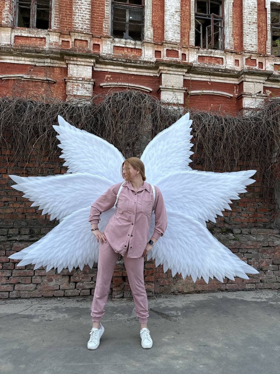 May include: A person wearing a pink shirt and pants stands in front of a brick wall with large white angel wings behind them.