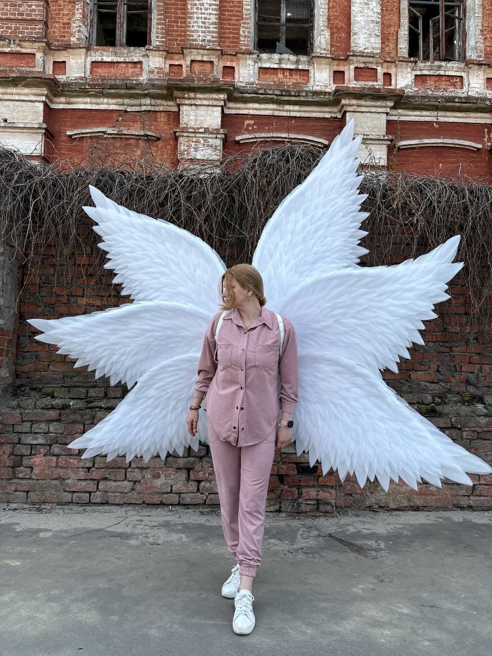 May include: A person wearing a pink shirt and matching pants stands in front of a brick wall with large white angel wings behind them.