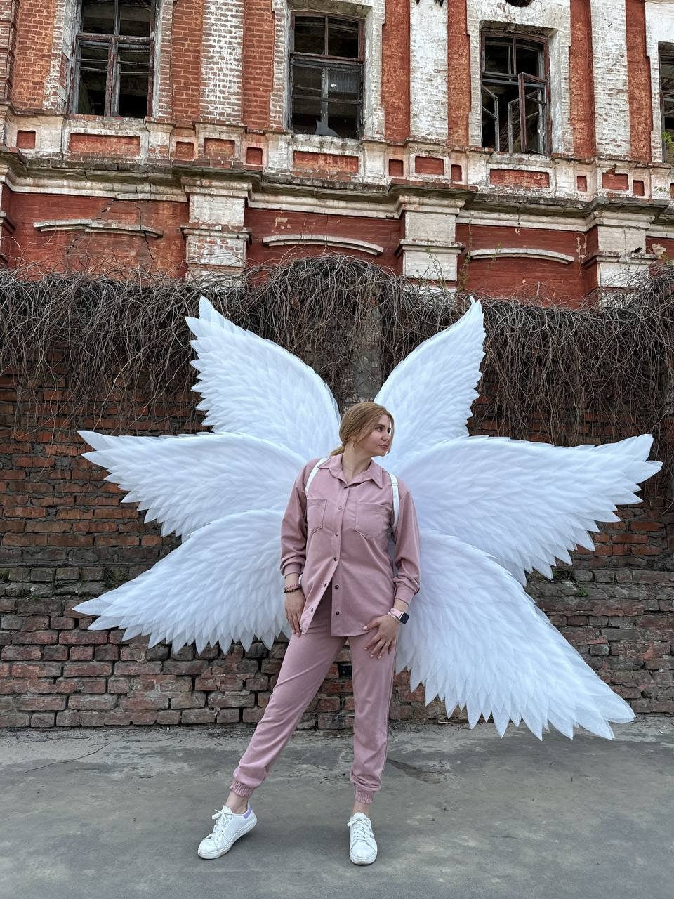 May include: A person wearing a pink jumpsuit and white sneakers stands in front of a brick building with large white angel wings behind them.