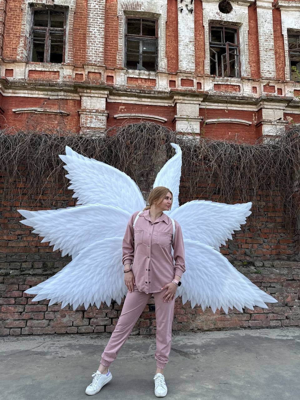 May include: A person wearing a pink outfit with white angel wings standing in front of a brick building.