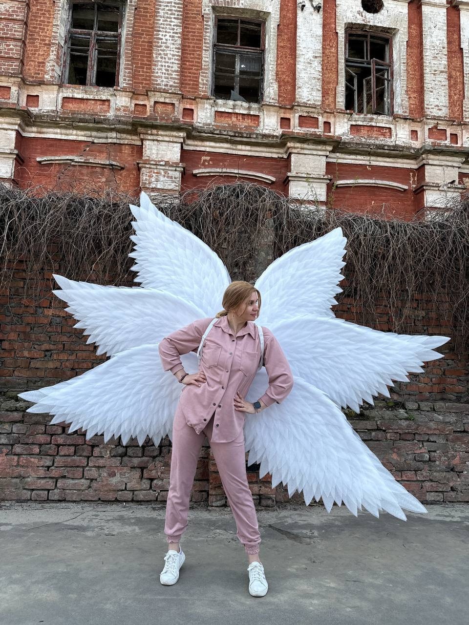 May include: A person wearing a pink jumpsuit and white sneakers stands in front of a brick building with large white angel wings.