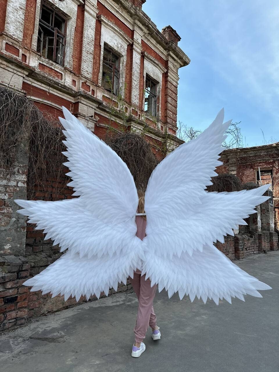 May include: A person wearing a pair of large, white angel wings. The wings are made of feathers and are very detailed. The person is standing in front of an old brick building.